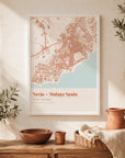 A white-framed location map print of Nerja, Malaga, Spain, hanging on a whitewashed wall in a terracotta and cream palette — styled with olive branches, a woven basket, and a terracotta jug, with a coastal Spanish village visible through an archway