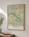 An oak-framed route map print hanging on a wall at an angle, showing a red route line traced across a detailed street map for the Manchester Marathon, styled with a plant and stacked books on a wooden console table