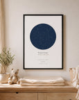 A framed navy star map print hanging above a wooden dresser in a nursery, styled with a potted olive tree, a crochet teddy bear, and folded muslin blankets