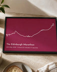 Personalised route trace print, in a black frame, landscape, with a white route line on a deep raspberry background above a title, date and finish time, shown on a kitchen table.