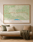 An oak-framed landscape route map print showing the Sussex Diamond Way trail traced in orange across a detailed green countryside map, hanging above a cream sofa styled with olive and tan cushions, a woven throw, walking boots, and hiking binoculars.