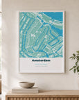 A white-framed location map print hanging above a wooden sideboard, showing a teal and sage green street map of Amsterdam's canals — styled with a stone bowl on stacked books and a potted plant.