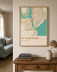An oak-framed location map print hanging in a hallway above a console table, showing a coastal town map in sage green, cream, and teal — styled with stacked books, a dried flower arrangement, and a blue sofa visible in the background