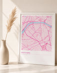 A white-framed location map print leaning against a wall, showing a hot pink and pale pink street map with a heart-shaped location marker, styled with a ceramic vase and dried pampas grass on a marble surface