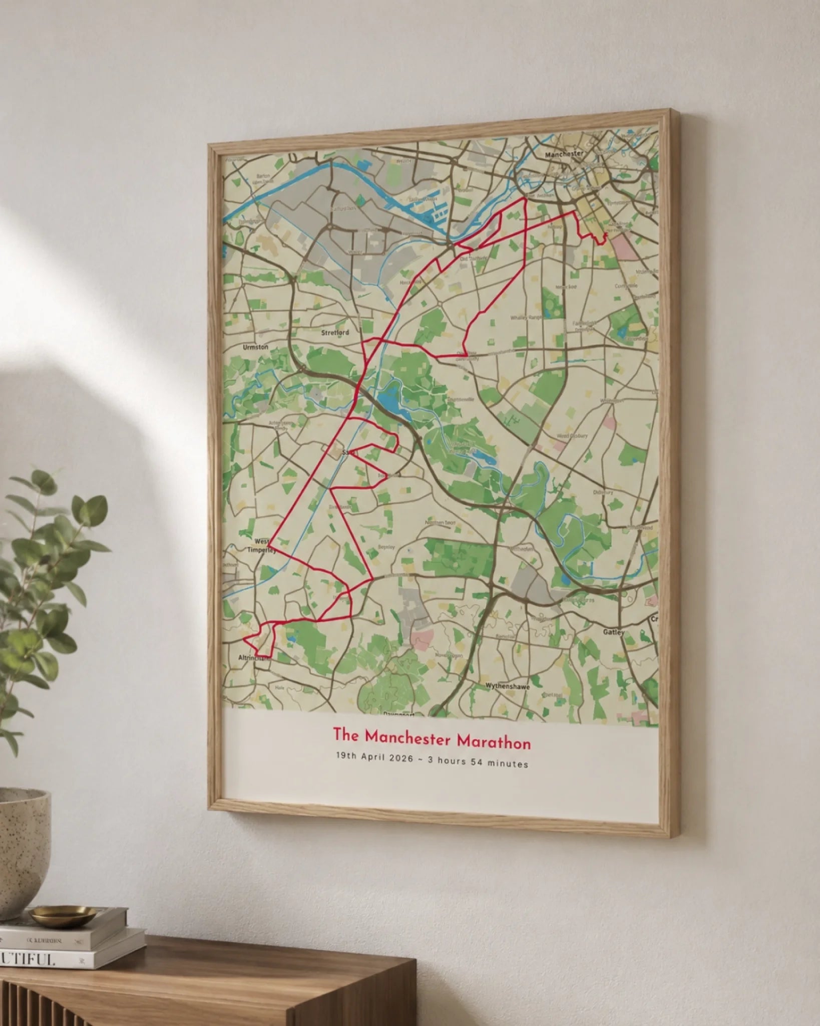 An oak-framed route map print hanging on a wall at an angle, showing a red route line traced across a detailed street map for the Manchester Marathon, styled with a plant and stacked books on a wooden console table