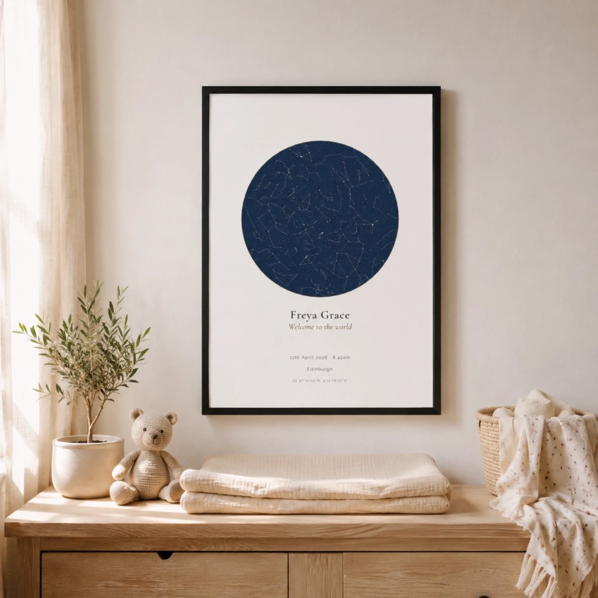A framed navy star map print hanging above a wooden dresser in a nursery, styled with a potted olive tree, a crochet teddy bear, and folded muslin blankets