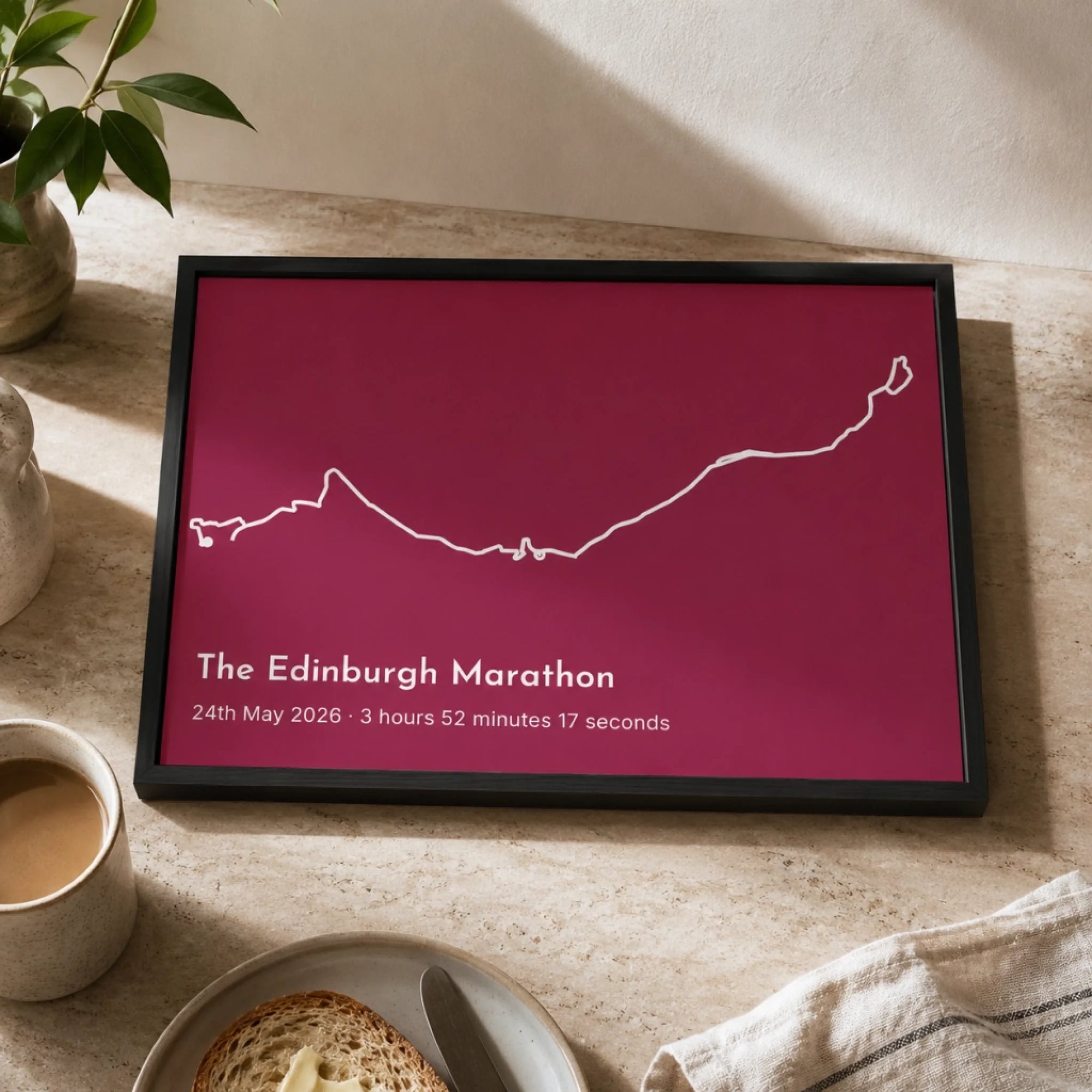 A framed personalized marathon print featuring a white outline map of The Edinburgh Marathon on a deep magenta background, styled on a light stone tabletop with a mug of coffee, toast, and a plant nearby.