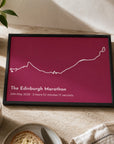 A framed personalized marathon print featuring a white outline map of The Edinburgh Marathon on a deep magenta background, styled on a light stone tabletop with a mug of coffee, toast, and a plant nearby.
