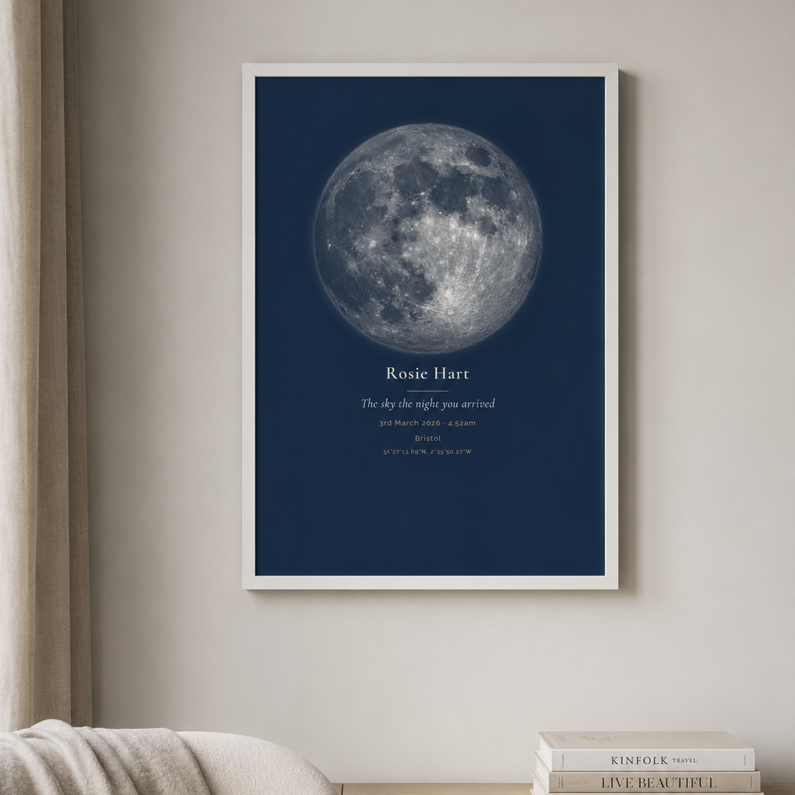 A white-framed moon phase print in a navy colourway, showing a detailed full moon against a dark blue sky, hanging above a stack of interior design books beside a curtained window and a throw blanket