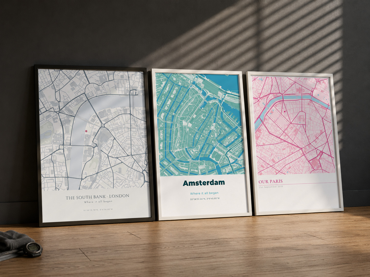 Three framed location map prints leaning against a wall — grey, teal, and pink street map styles of London, Amsterdam, and Paris