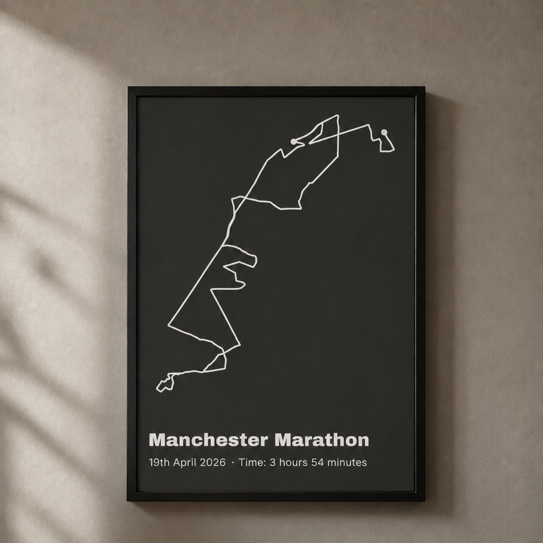 A black-framed route map print in a dark charcoal colourway, hanging on a sunlit wall with dappled shadow — a cream route line traces the Manchester Marathon shape, with the event name and time printed below