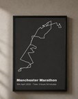 A black-framed route map print in a dark charcoal colourway, hanging on a sunlit wall with dappled shadow — a cream route line traces the Manchester Marathon shape, with the event name and time printed below